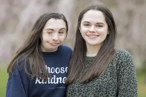 Twin sisters, including one wearing a blue sweatshirt that says Choose Kindness, are standing in front of a blurred background.