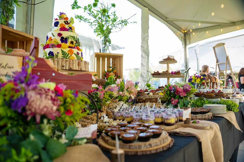 table filled with various food platters and natural burlap and flower decor, highlighted by a white cake adorned with colorful wildflowers Marywood University/Gourmet Dining Capture Third Consecutive National Dining Award