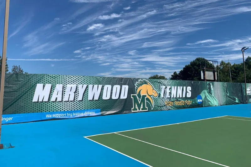 A green tennis court surrounded by light blue with a Marywood banner covering the fence that engulfs the court