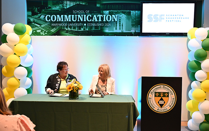 two women signing a document against a backlit wall and green, yellow, and white balloon decorations with a wall banner that says School of Communication and Scranton Shakespeare Festival