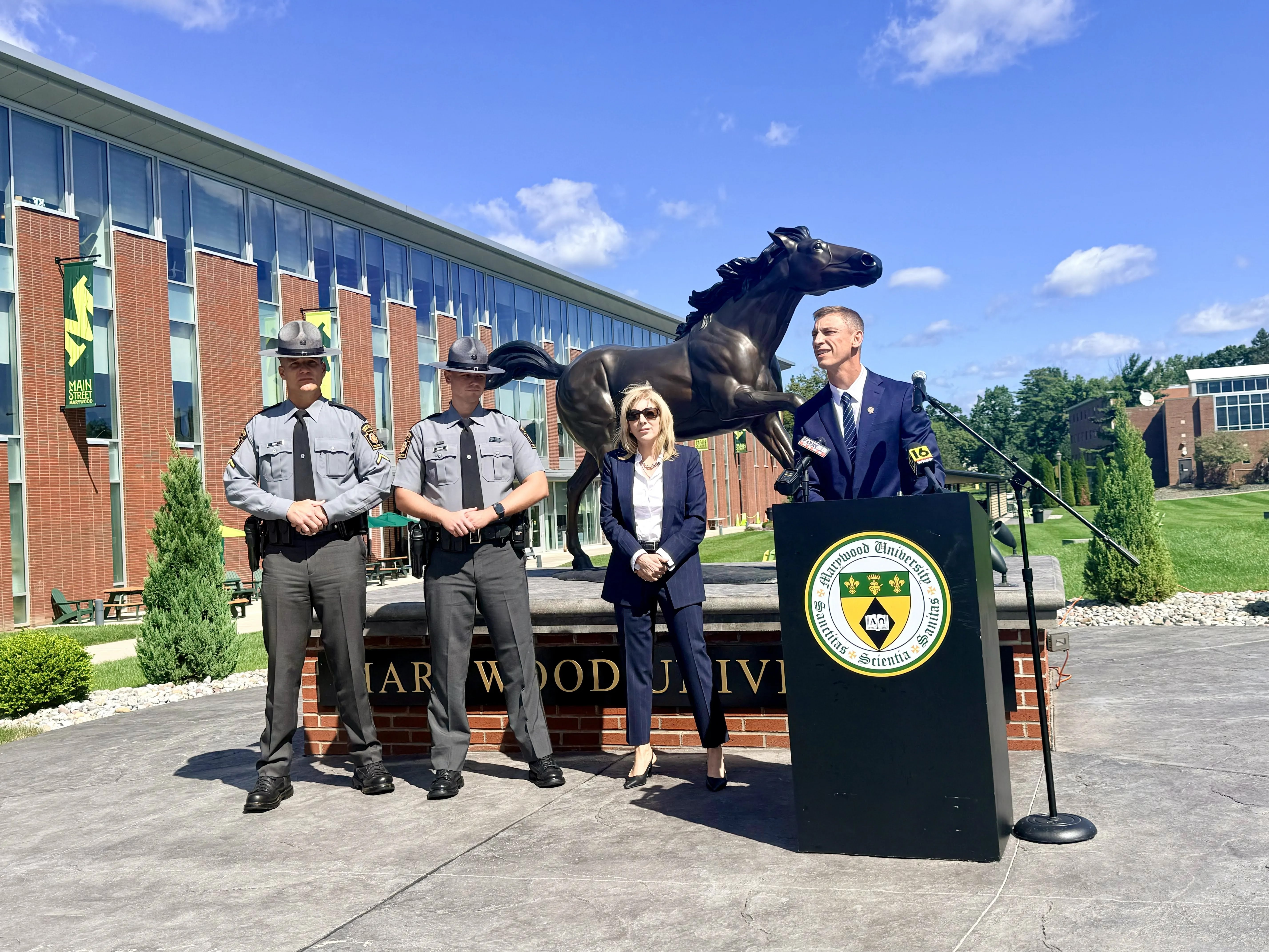 Two Pennsylvania State Troopers, a woman in a navy suit and a man in a navy suit behind a podium outside in front of a bronze horse statue and blue sky.