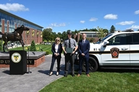 A woman in a navy suit and a man in a navy suit with a state trooper in the middle near a bronze horse statue, a state police SUV, and blue sky background. Marywood University and Pennsylvania State Police Academy Partnership Celebrate Milestone of First Graduate