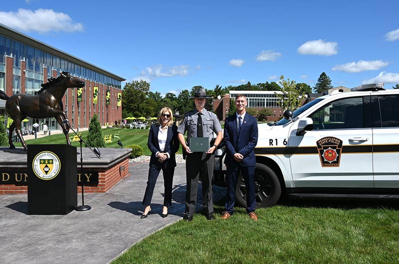 A woman in a navy suit and a man in a navy suit with a state trooper in the middle near a bronze horse statue, a state police SUV, and blue sky background.