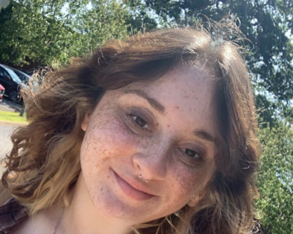 A smiling woman with curly hair, freckles, and a nose ring, outside on a sunny day with trees in the background.