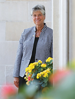 Sister Mary leaning on a wall smiling along with yellow flowers in front of her