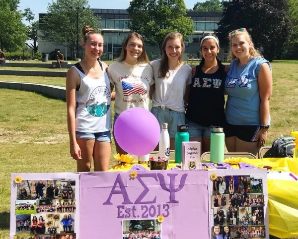 Five women standing outside and smiling behind a table with Alpha Sigma Psi poster and a purple balloon